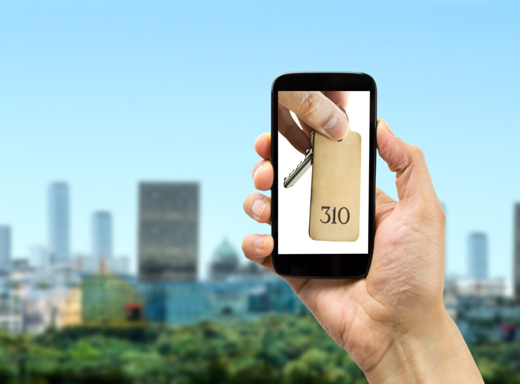 Hand holding a smartphone with a picture of keys of hotel room with a cityscape on the background.
